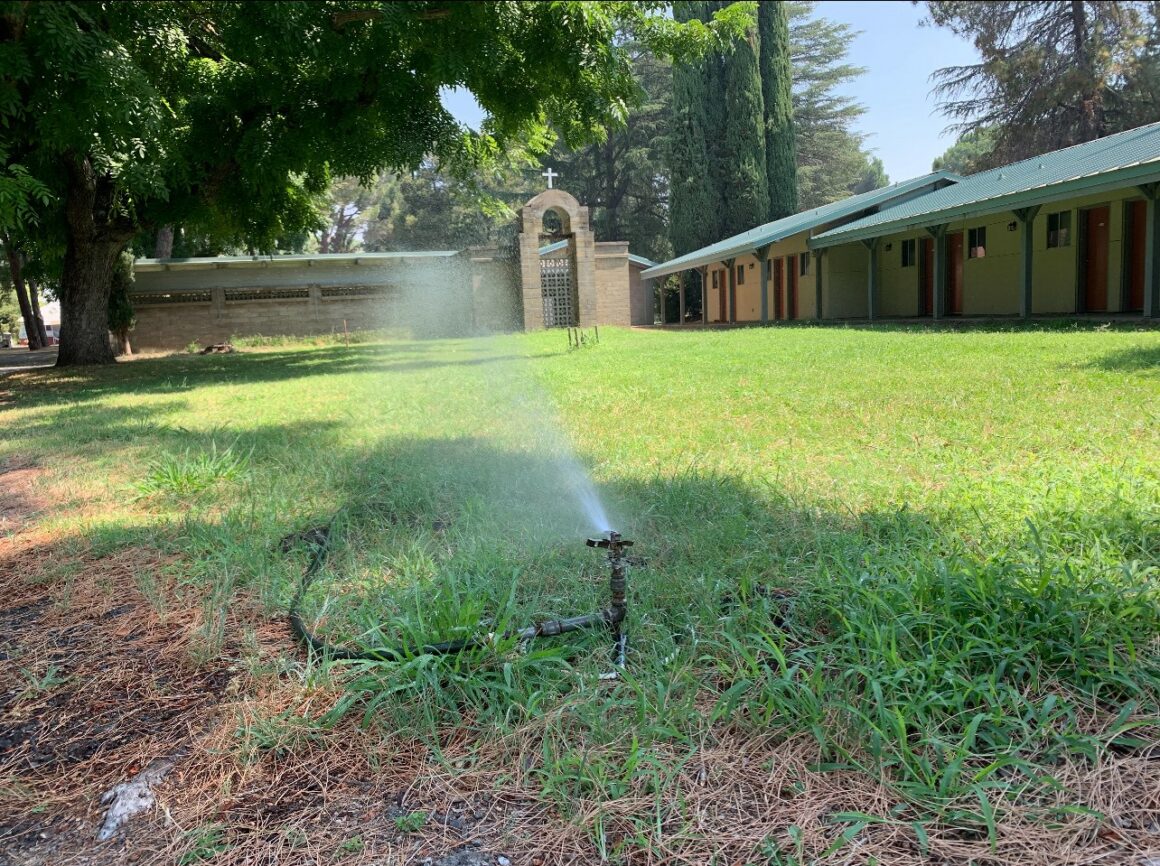 a sprinkler waters a green lawn in from of monastery buildings